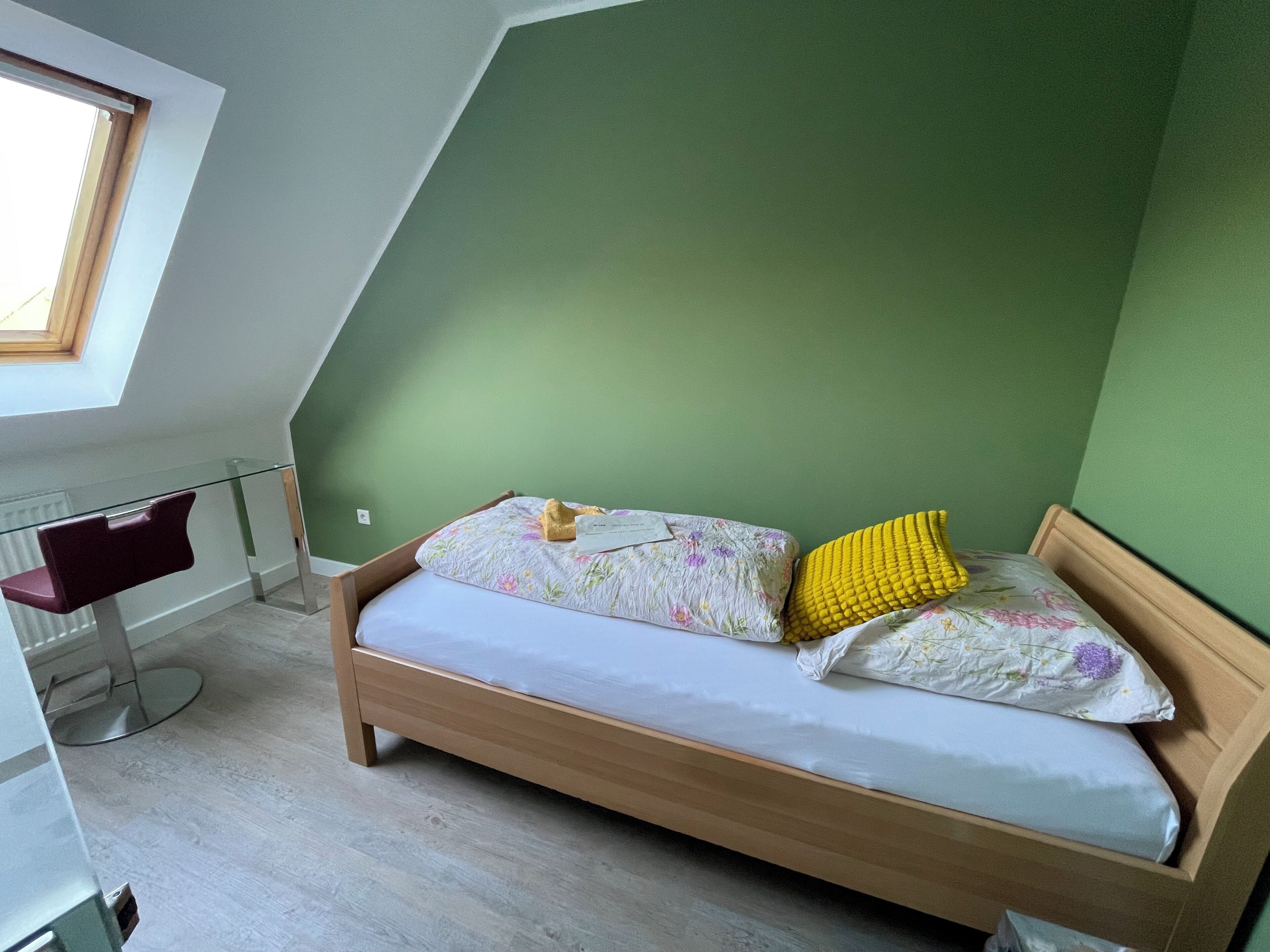 A room with a sloped ceiling, a bed with floral bedding, a yellow pillow, and a desk with a chair.