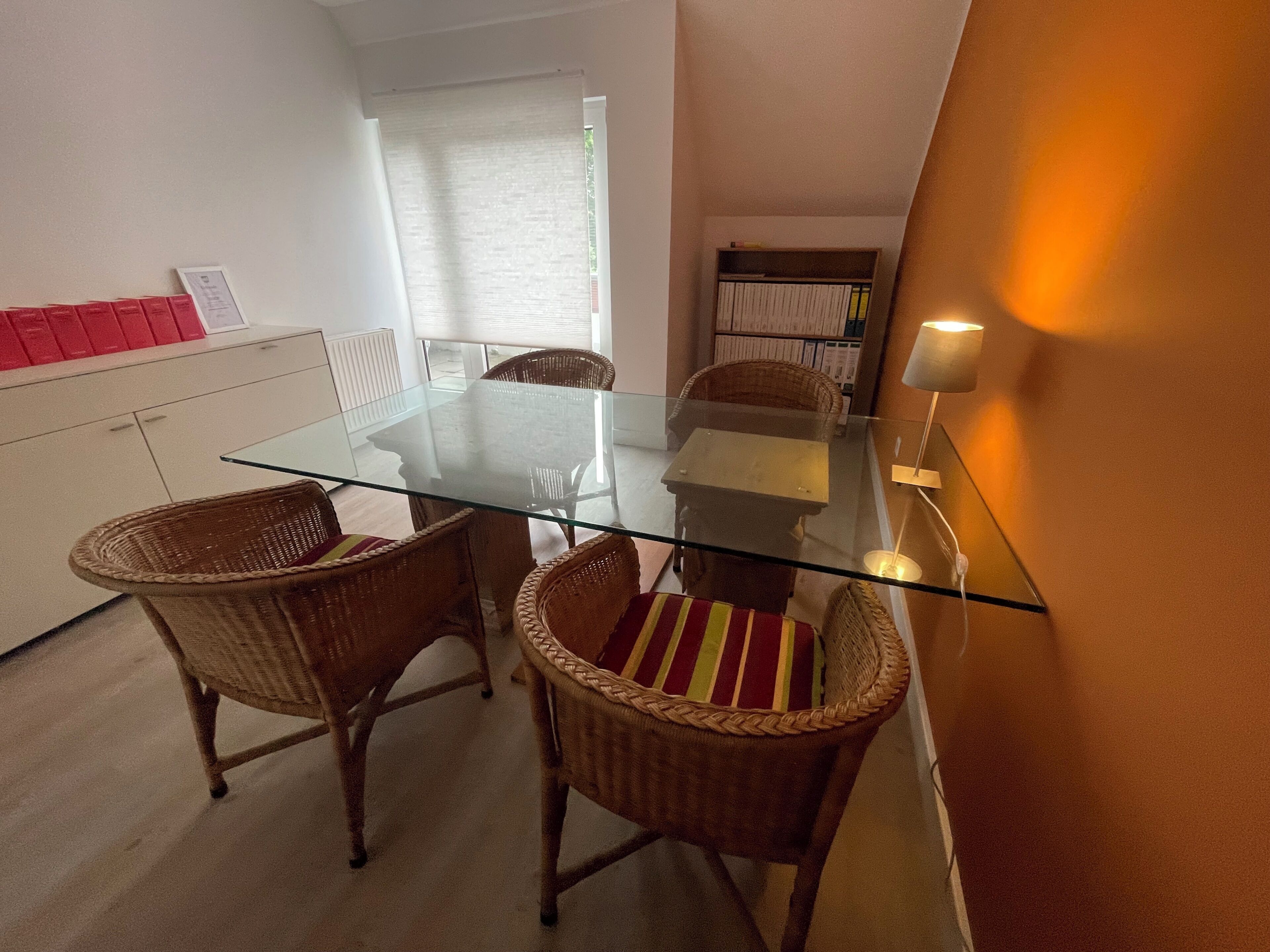 Un comedor con una mesa de vidrio y sillas de mimbre, un sofá blanco con cojines rojos, y una estantería al fondo.