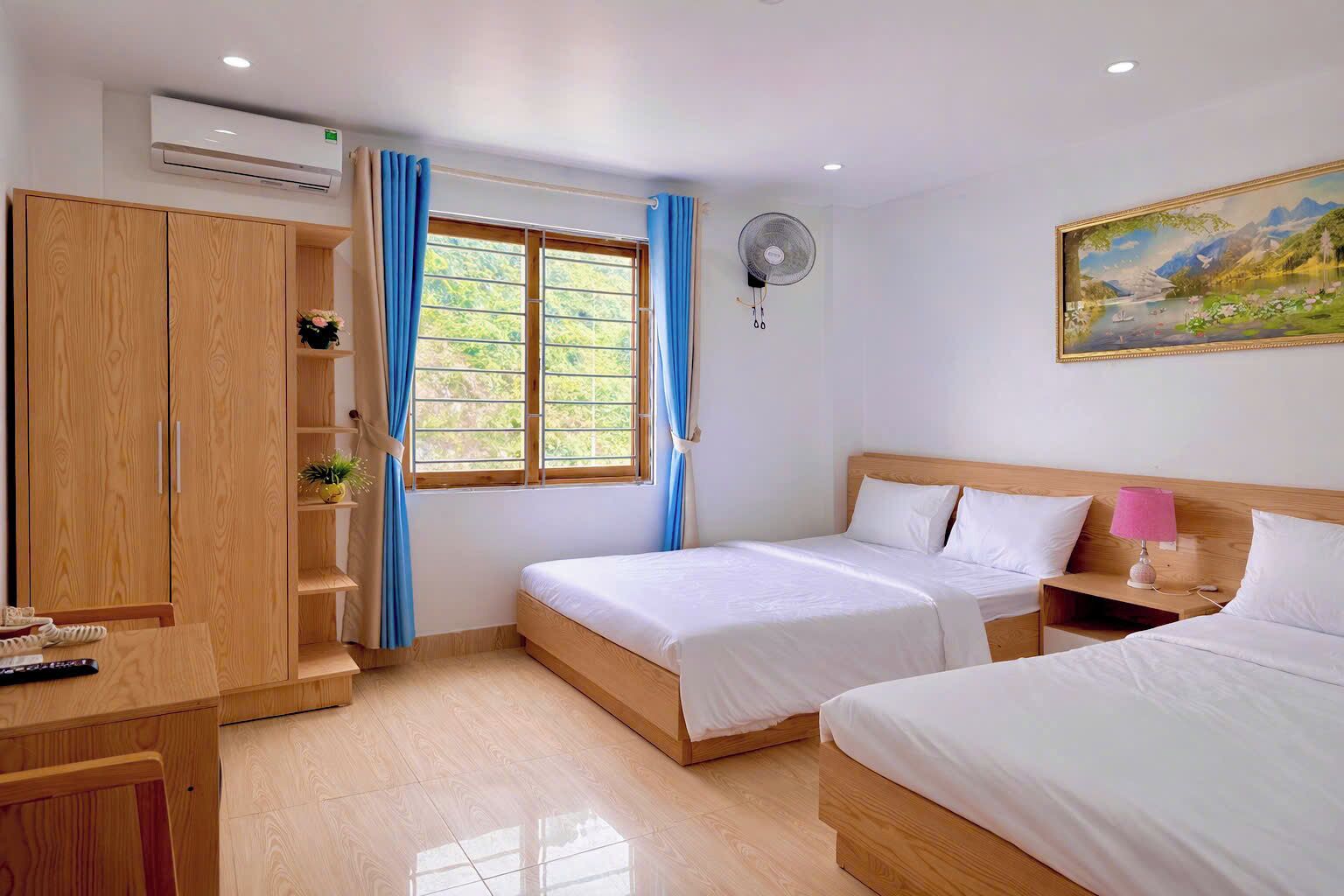 Een hotelkamer met twee bedden, een houten kledingkast, een bureau met stoel, een raam met gordijnen, een ventilator aan de muur en een schilderij.