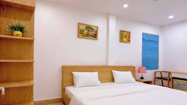Un dormitorio con cama de madera, ropa de cama blanca, una estantería de madera con una planta y dos cuadros enmarcados en la pared.