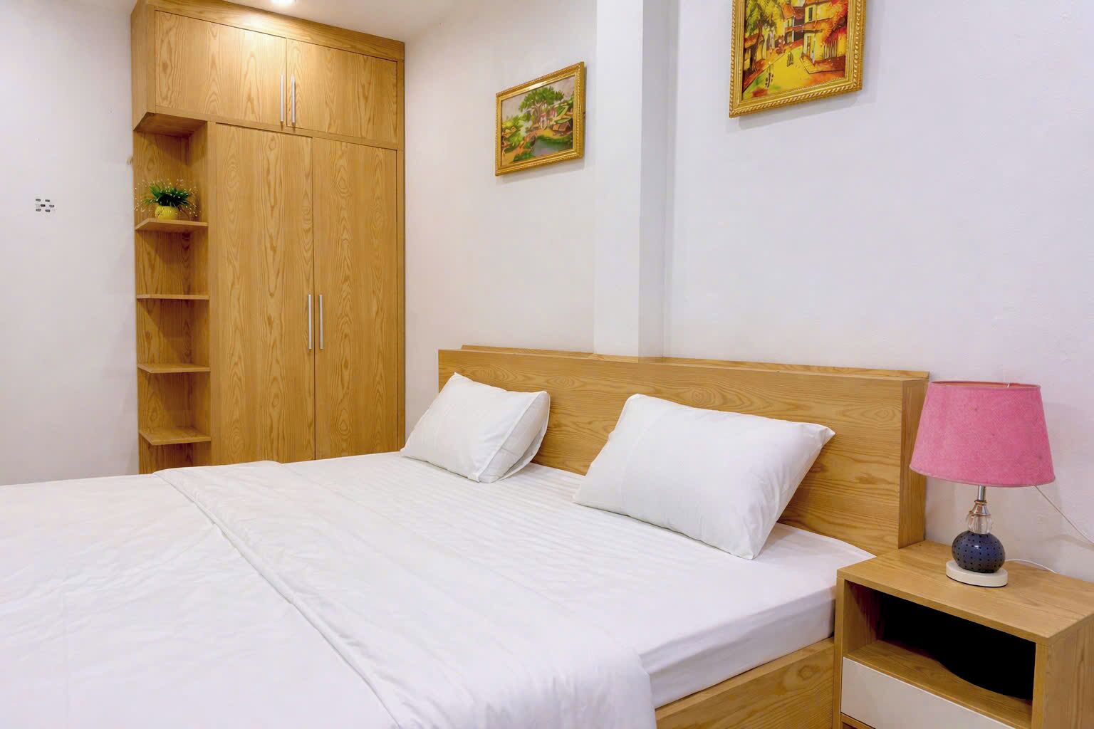 A bedroom with a bed, wooden wardrobe, two paintings, and a bedside lamp.