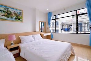 Habitación de hotel con cama, mesita de noche, lámpara, un cuadro, ventana con cortinas y vistas a edificios.