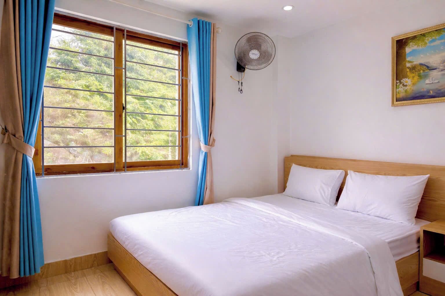 A bedroom with a bed, a window with curtains, a wall-mounted fan, and a painting.