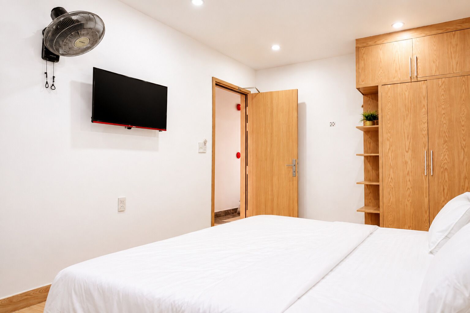 Un dormitorio con una cama, un televisor montado en la pared, un armario de madera y una puerta con picaporte rojo.