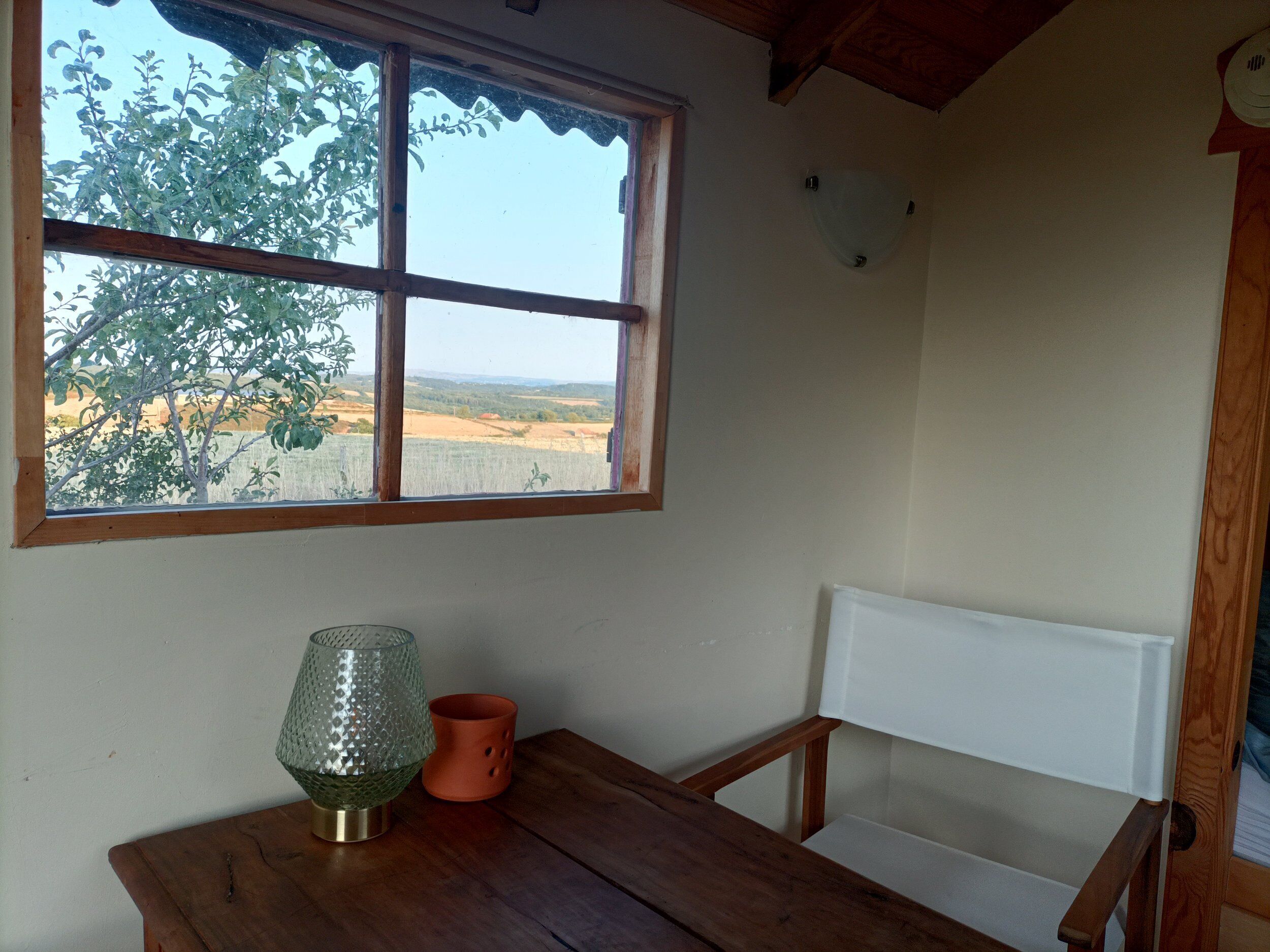 Una habitación con una mesa de madera, una lámpara verde con textura, un florero color cobre y vista a un paisaje a través de una ventana.