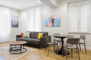 A modern living room with a grey sofa, a round coffee table, and a dining table with chairs. There are two paintings on the walls, a window with white curtains, and a wooden floor. - Casa e Appartamento Vacanze Gallese 4 (Roma)