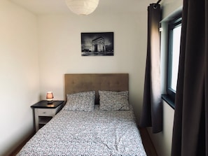 Ein Schlafzimmer mit einem Bett, einem Nachttisch, einer Lampe, einem Fenster mit Vorhängen und einem Bild an der Wand.