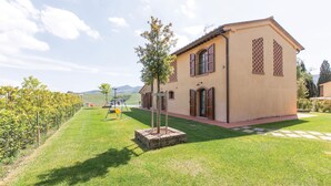 A single-story building with a tiled roof, a small garden with a swing set, and a vineyard in the background. - Nice home in Lajatico with private swimming pool, can be inside or outside (Lajatico)