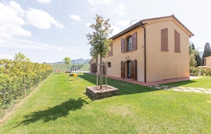 Ein eingeschossiges Gebäude mit Ziegeldach, ein kleiner Garten mit Schaukel und ein Weinberg im Hintergrund.