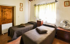 A room with two single beds, a wooden door, a window with a curtain, a bedside table, and a standing fan. - Nice home in Caprese Michelangelo (Caprese Michelangelo)