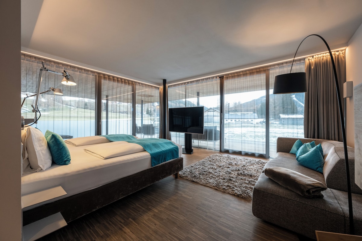 A modern hotel room with a large bed, a sofa, a TV, and a view of a snowy landscape.