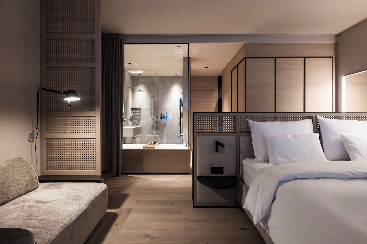 A modern hotel room with a bed, a seating area, and a shower.
