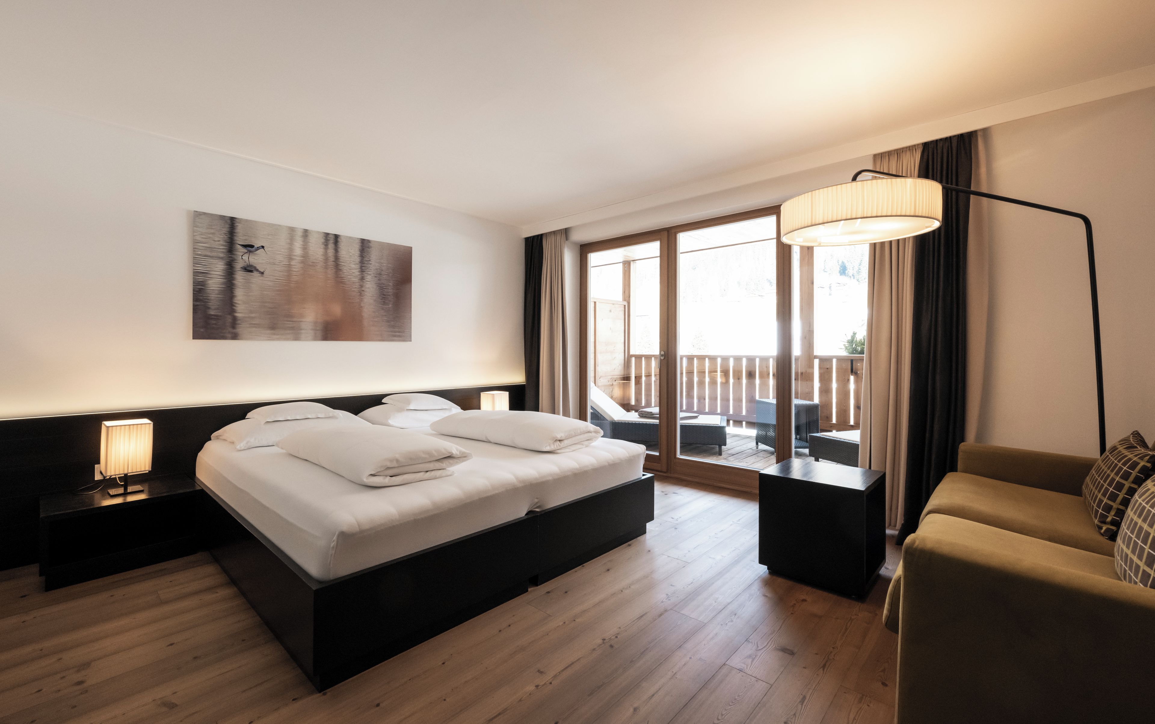 Une chambre d’hôtel comprenant un lit, une table de chevet, une lampe, un tableau, un balcon avec une table et un canapé.