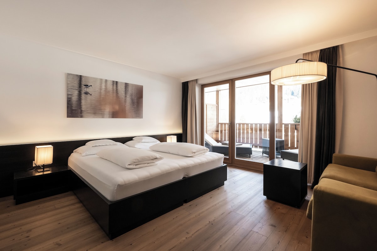 A hotel room with a bed, a nightstand, a lamp, a painting, a balcony with a table, and a sofa.