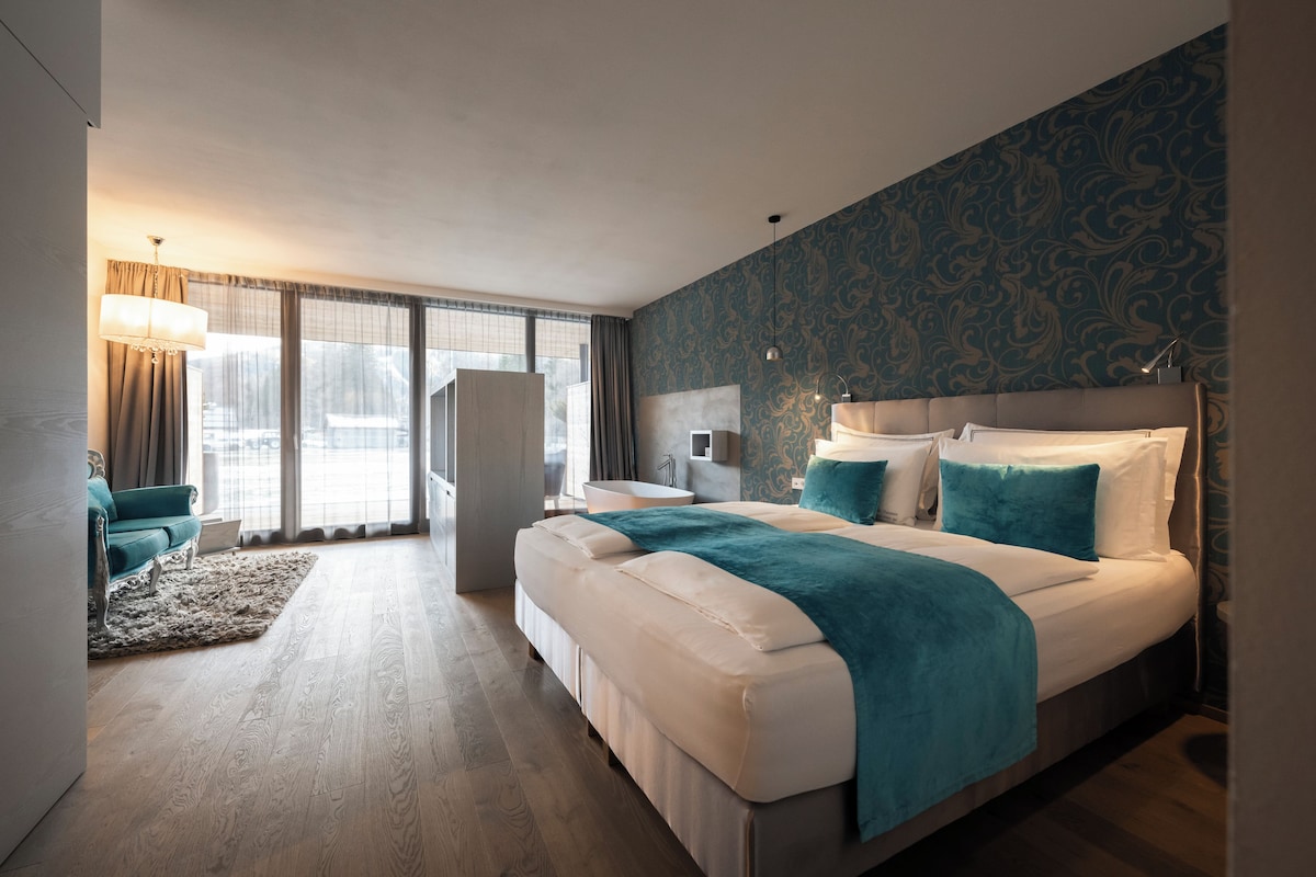 A modern hotel room with a large bed, a sitting area with a sofa and armchair, a small desk, and a view through the window.