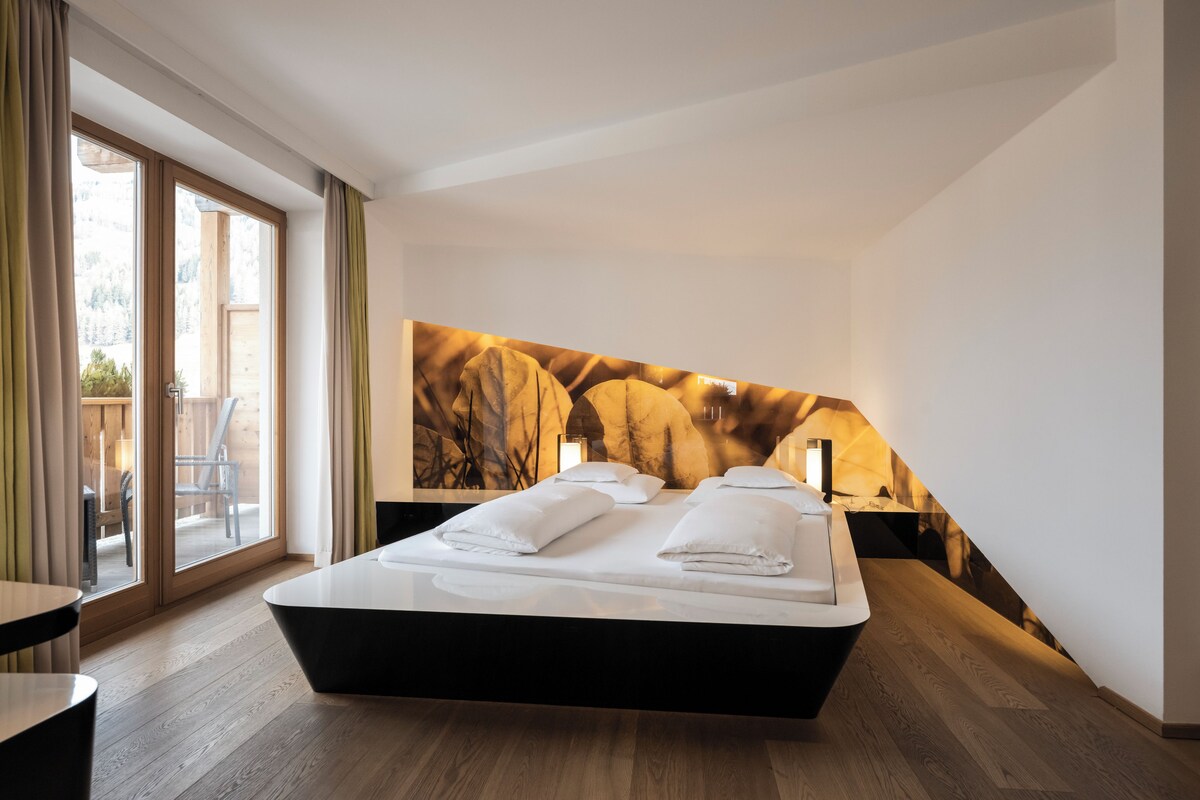 A modern bedroom with a large bed, a view of a mountainous landscape through a window, and a wooden floor.