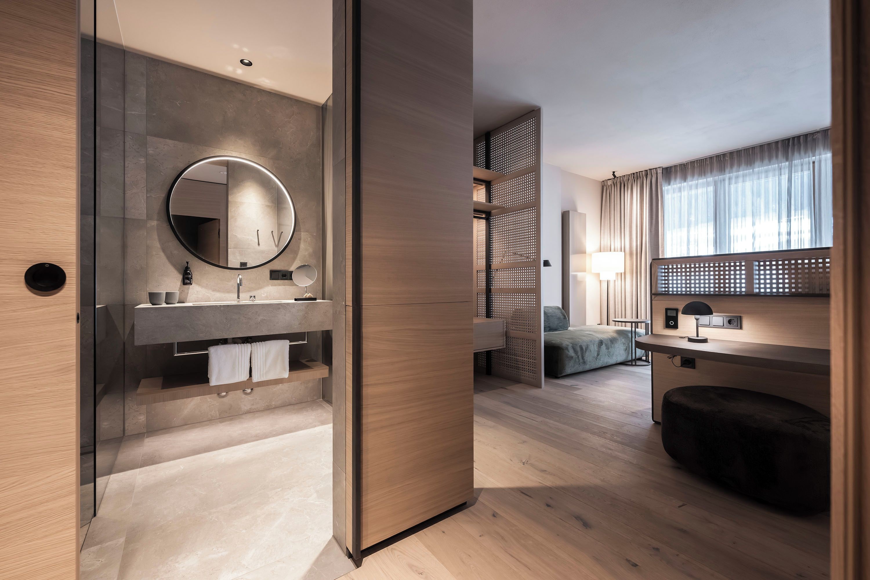 A modern hotel room with a wooden floor, a round mirror, a wooden cabinet, a bed, and a desk with a lamp.