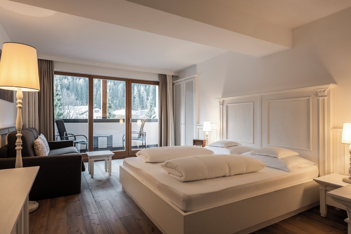 A hotel room with a large bed, a sitting area with a chair and a table, and a balcony with a view of trees.