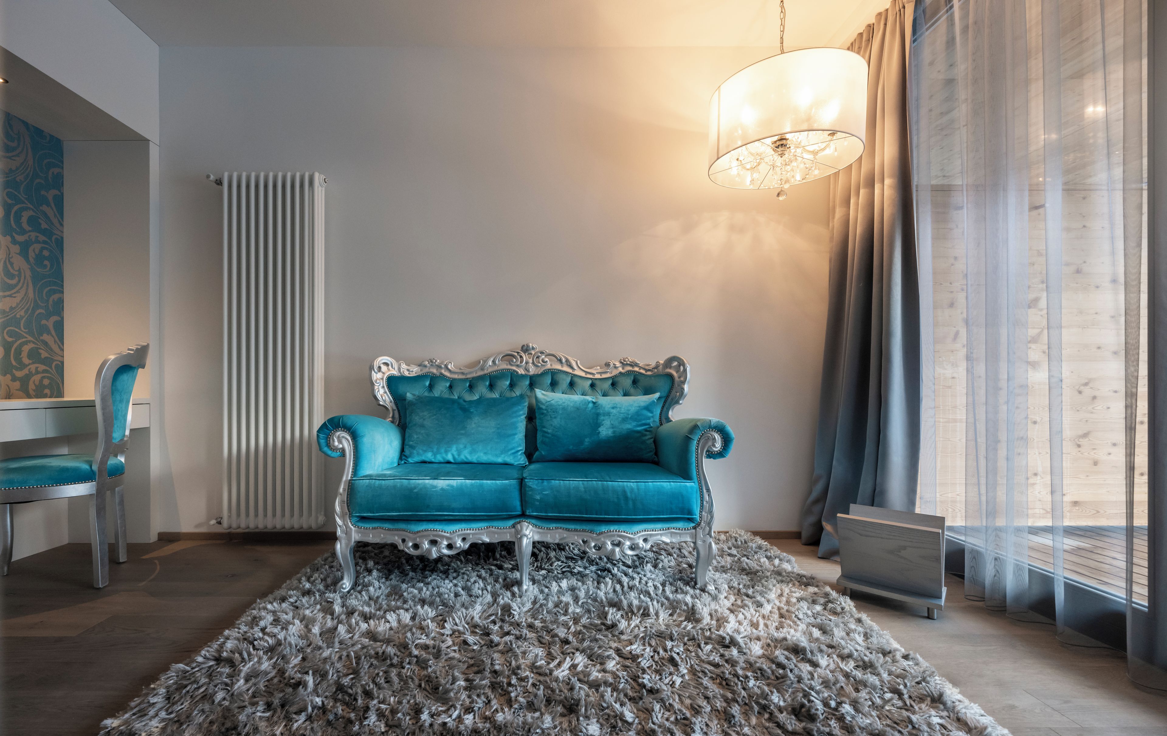 A room with a teal sofa, a patterned rug, a radiator, a chair, and a hanging light fixture.