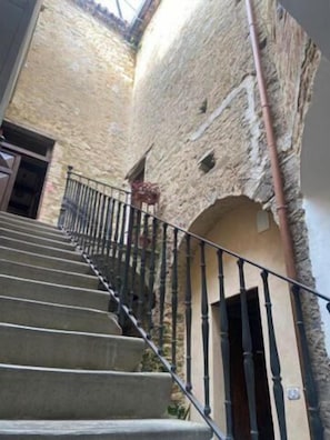 Una escalera con barandilla metálica que lleva a un nivel superior, rodeada de muros de piedra y una puerta al final.