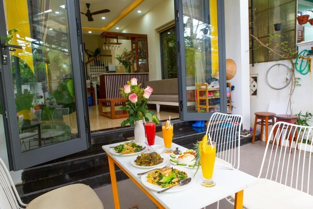 Un área de comedor al aire libre con una mesa lista para dos personas, un jarrón de flores y una variedad de platos y bebidas.