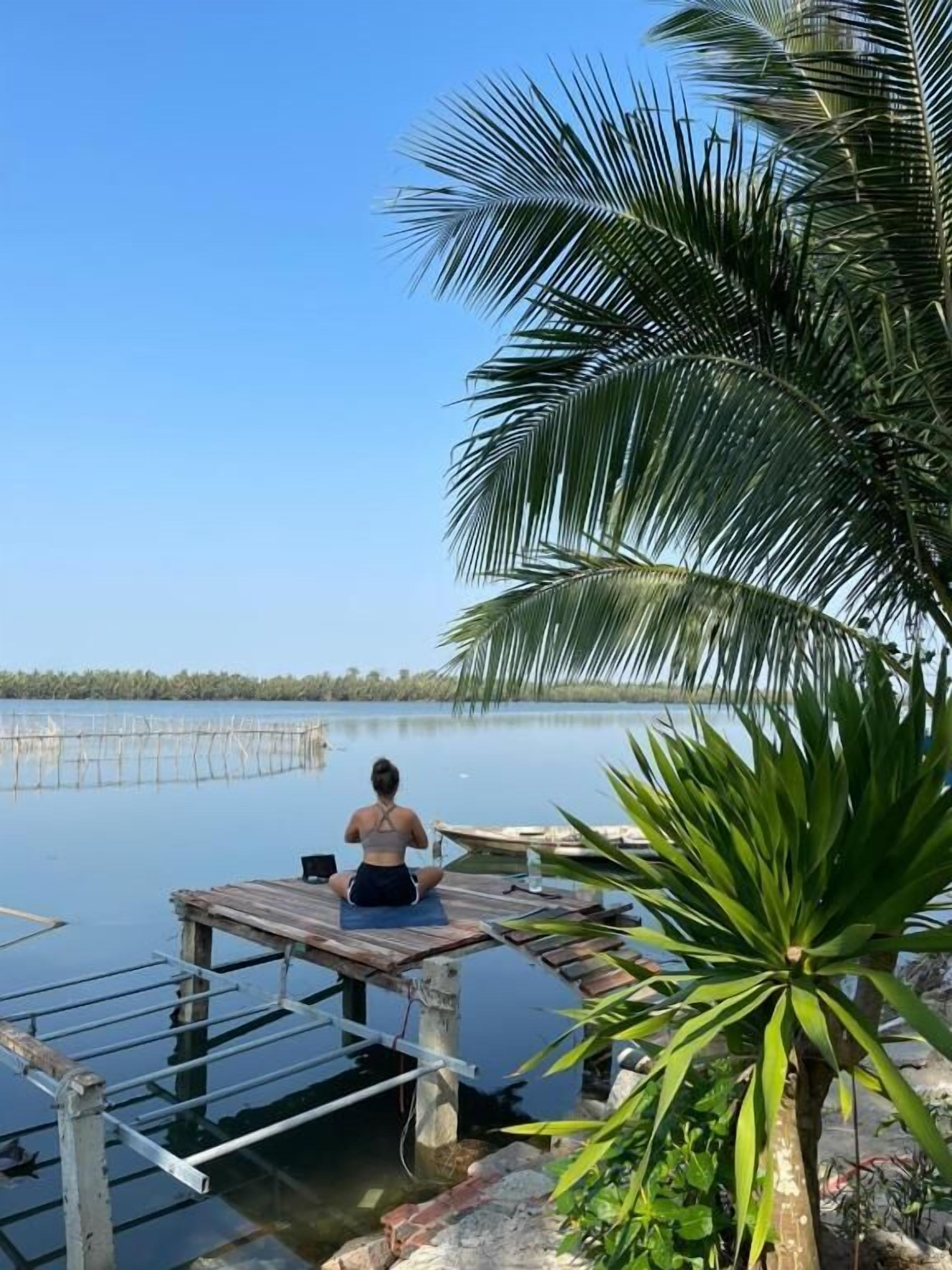 Una persona sentada sobre un muelle de madera que se extiende hacia aguas tranquilas, rodeada de palmeras y un cielo despejado.