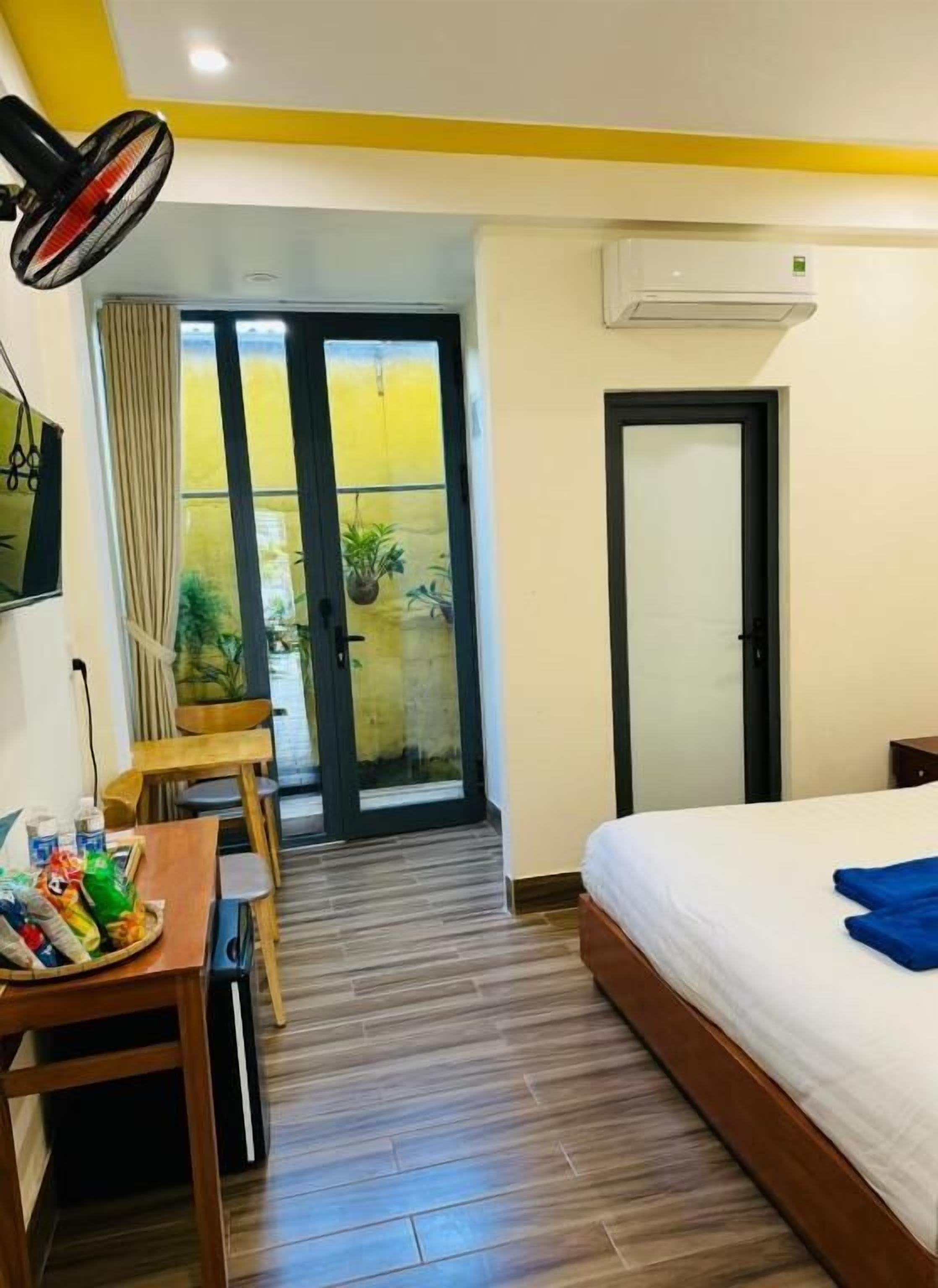 Habitación de hotel con cama, escritorio con aperitivos, ventilador y puerta corrediza de vidrio que da a un balcón con plantas.
