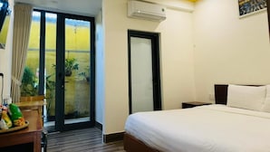 A hotel room with a bed, a desk with a fan, a window with curtains, an air conditioning unit, and a door.
