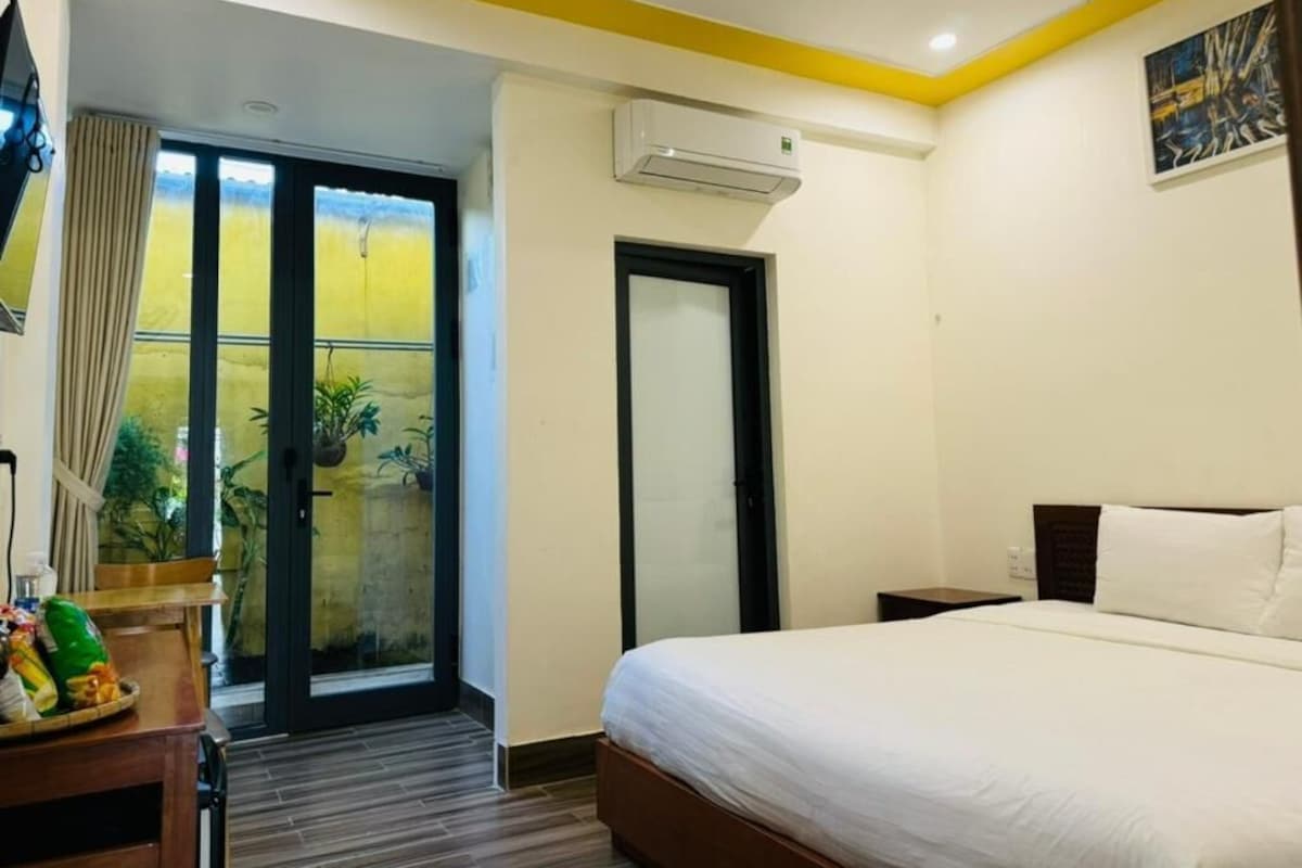 A hotel room with a bed, a desk with a fan, a window with curtains, an air conditioning unit, and a door.