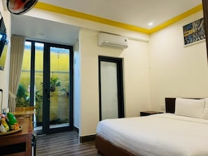 A hotel room with a bed, a desk with a fan, a window with curtains, an air conditioning unit, and a door. - Coral Riverside Homestay & Hostel (Da Nang)