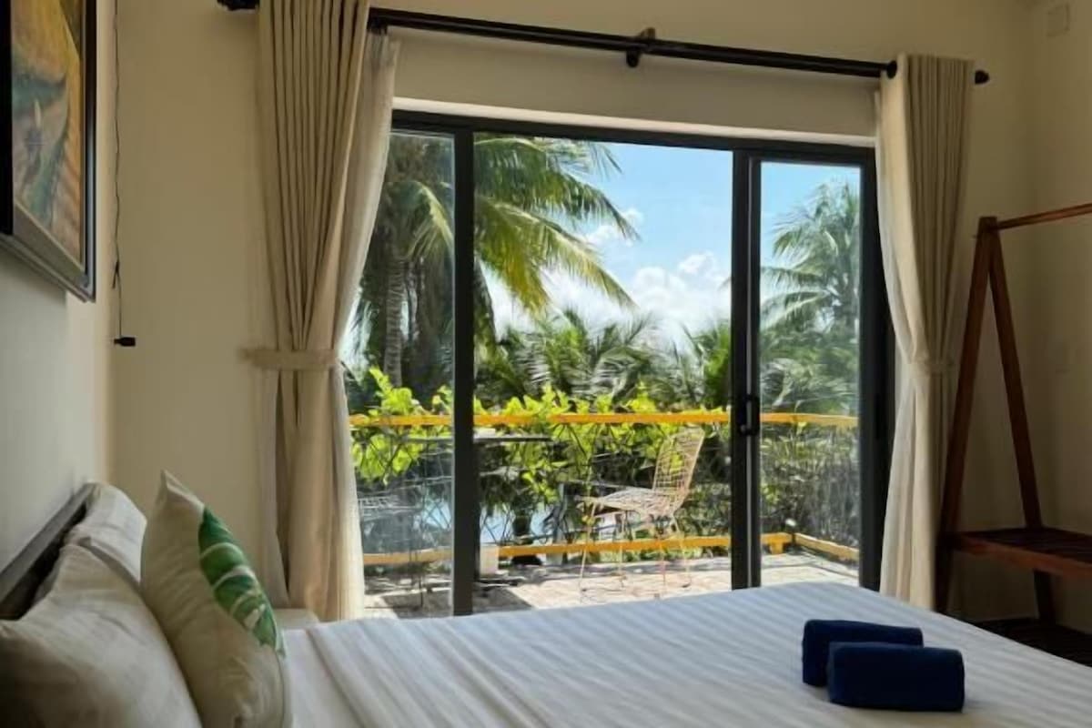 A hotel room with a bed, a view of a balcony with palm trees, and a swing chair.