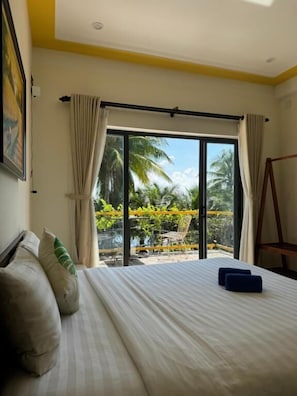 A hotel room with a bed, a view of a balcony with palm trees, and a swing chair. - CORAL RIVERSIDE HOMESTAY & HOSTEL (Hoi An Tay)
