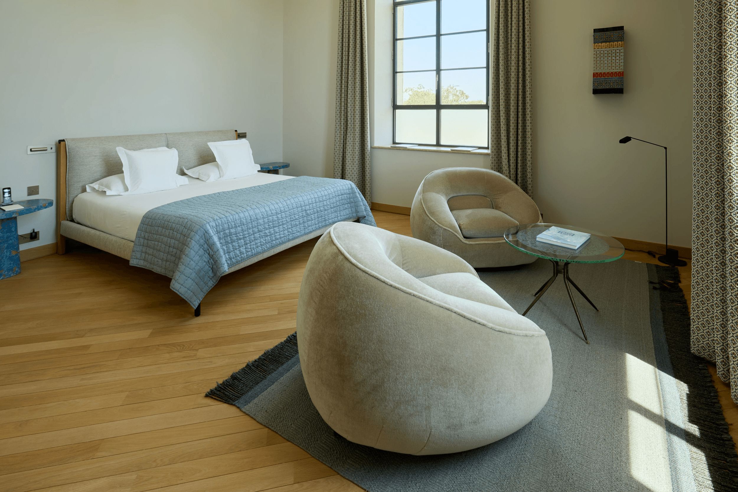 A modern bedroom with a bed, a round chair, a small glass table, and a window with curtains.