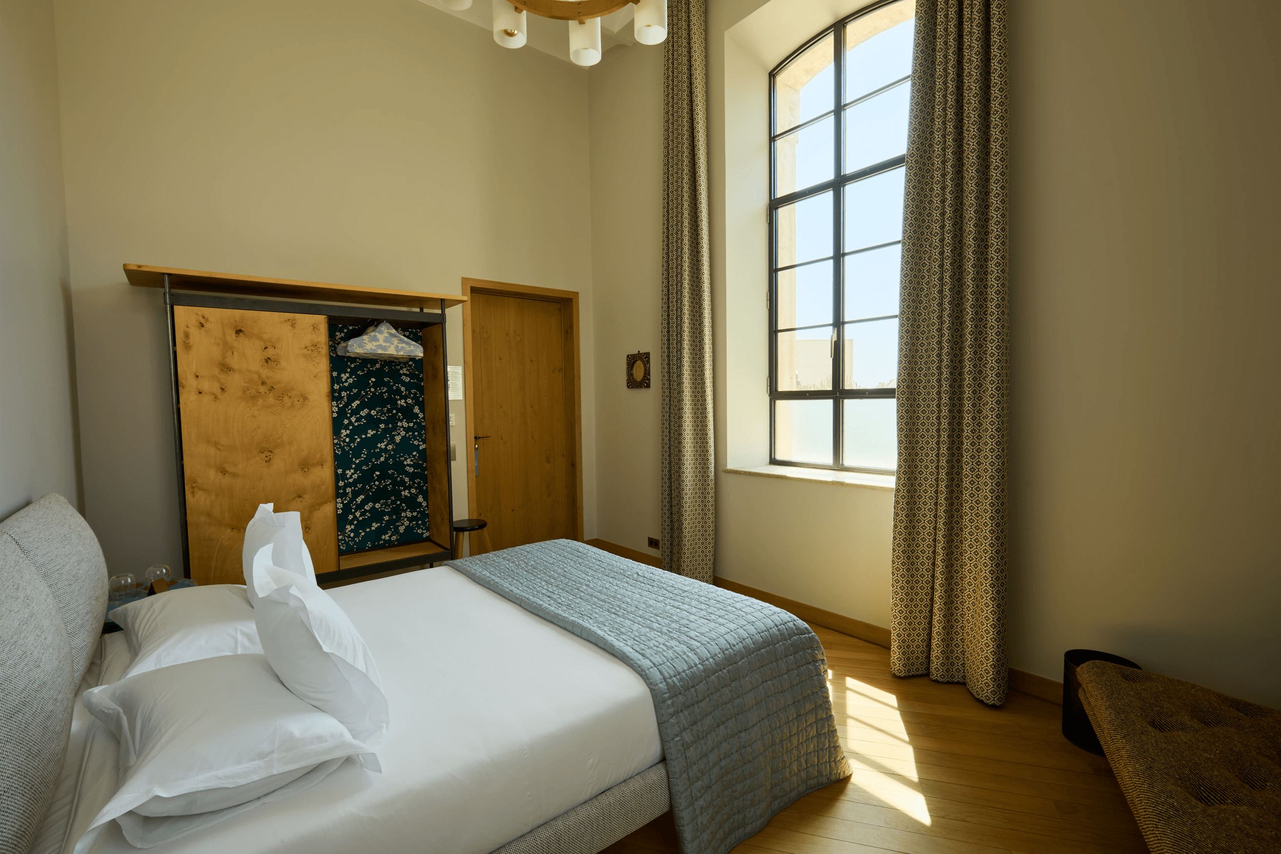 A bedroom with a bed, a wardrobe, a window with curtains, and a small stool.