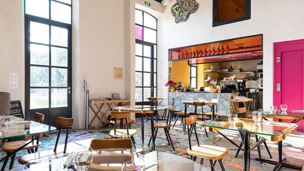 Un comedor amplio con varias mesas y sillas, ventanas grandes y una cocina visible a través de un umbral abierto.