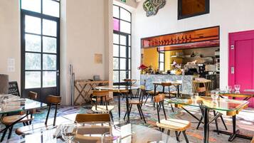 Un grand espace dédié à la salle à manger, avec plusieurs tables et chaises, de grandes fenêtres et une cuisine visible à travers une porte ouverte.