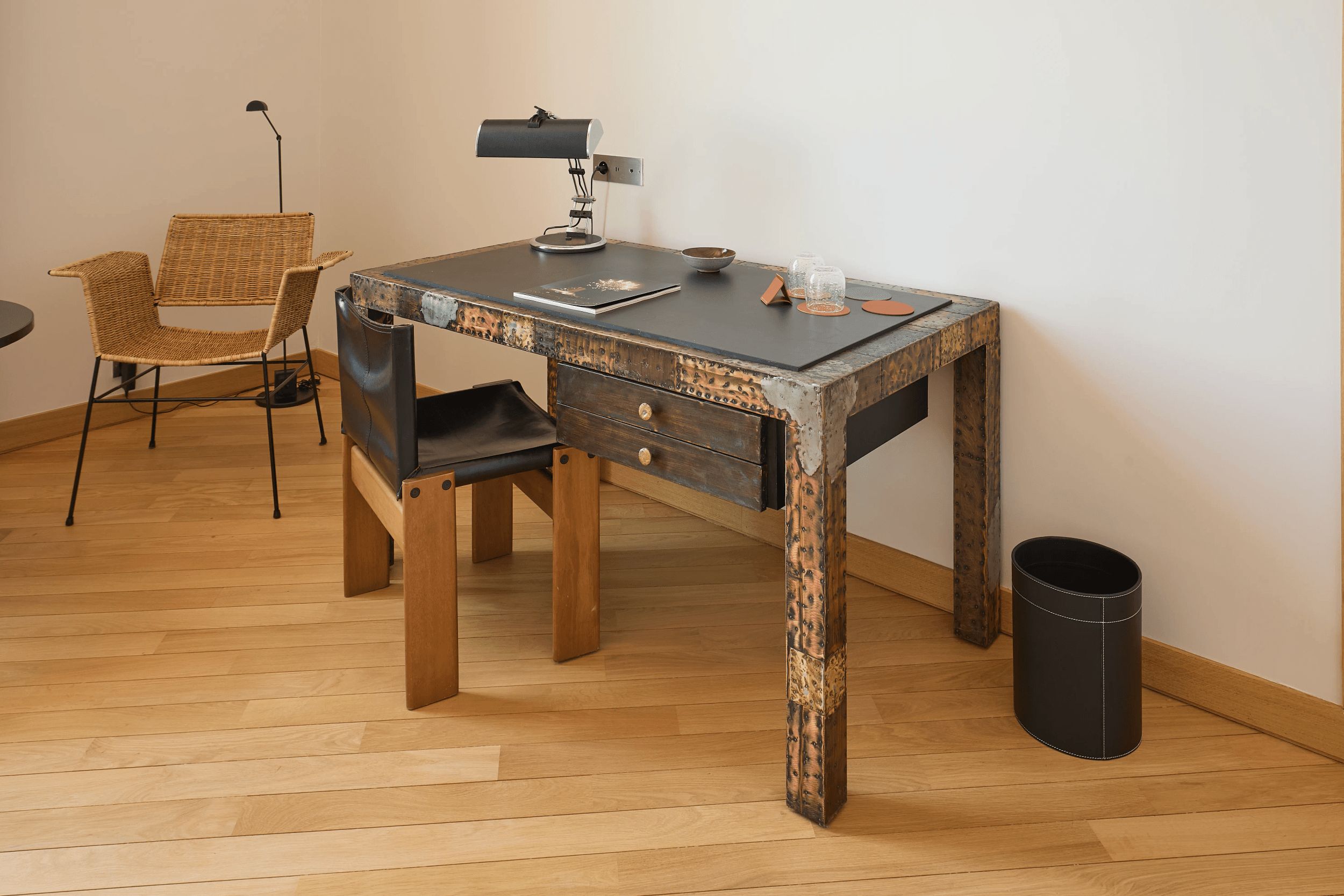 Un escritorio de madera con una lámpara metálica, una silla de mimbre y un cesto de basura.