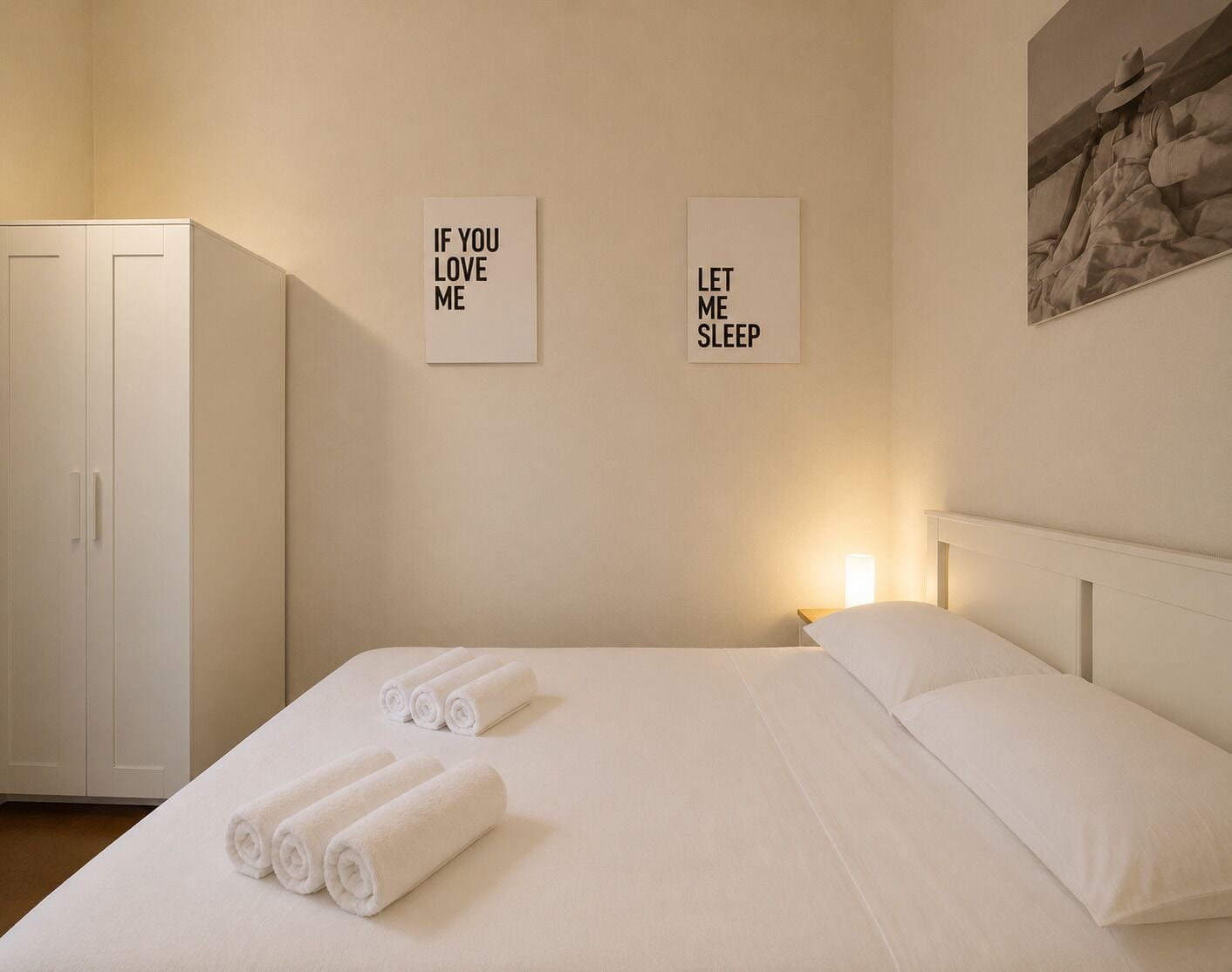 Hotelkamer met een bed, twee opgerolde handdoeken, een nachtkastje met een lamp, en twee decoratieve wandpanelen met tekst.