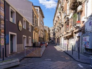 A narrow urban street with buildings on both sides, some with balconies and signage.