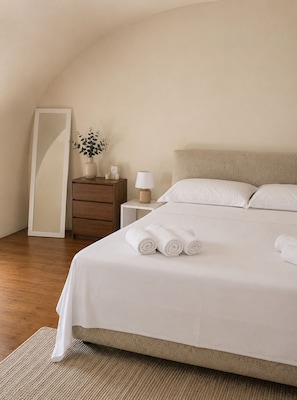 Ein ordentlich bezogenes Bett mit aufgerollten Handtüchern, ein Nachttisch mit Lampe, ein Beistelltisch mit Vase und Bilderrahmen sowie ein großer Spiegel an der Wand.