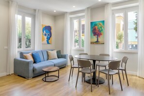 A modern living room with a grey sofa, a round dining table, and six chairs. There are two windows with white curtains, and two paintings are hung on the walls. - Casa e Appartamento Vacanze Gallese 5 (Roma)