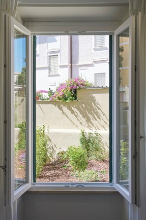 Ein Fenster mit Blick auf ein Gebäude, einen Garten mit verschiedenen Pflanzen und Blumen, und einen klaren Himmel.