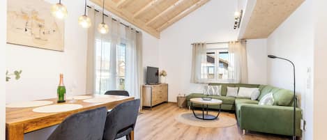 A spacious living area with a wooden ceiling, a dining table with chairs, a green sofa, and a floor lamp.