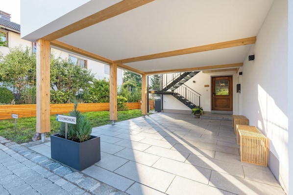 A covered outdoor area with a staircase leading to a door, a planter with greenery, and a bench.