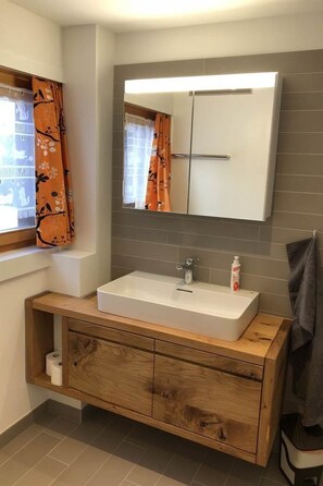 Un baño con lavamanos blanco, tocador de madera, paredes de cerámica gris y un espejo sobre el lavamanos.