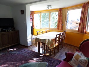 Uma sala de jantar com uma mesa redonda, cadeiras, uma televisão e uma janela com cortinas.