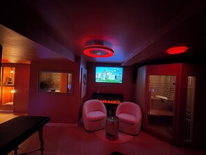 A room with a fireplace, two chairs, a bench, and a television displaying a cityscape.