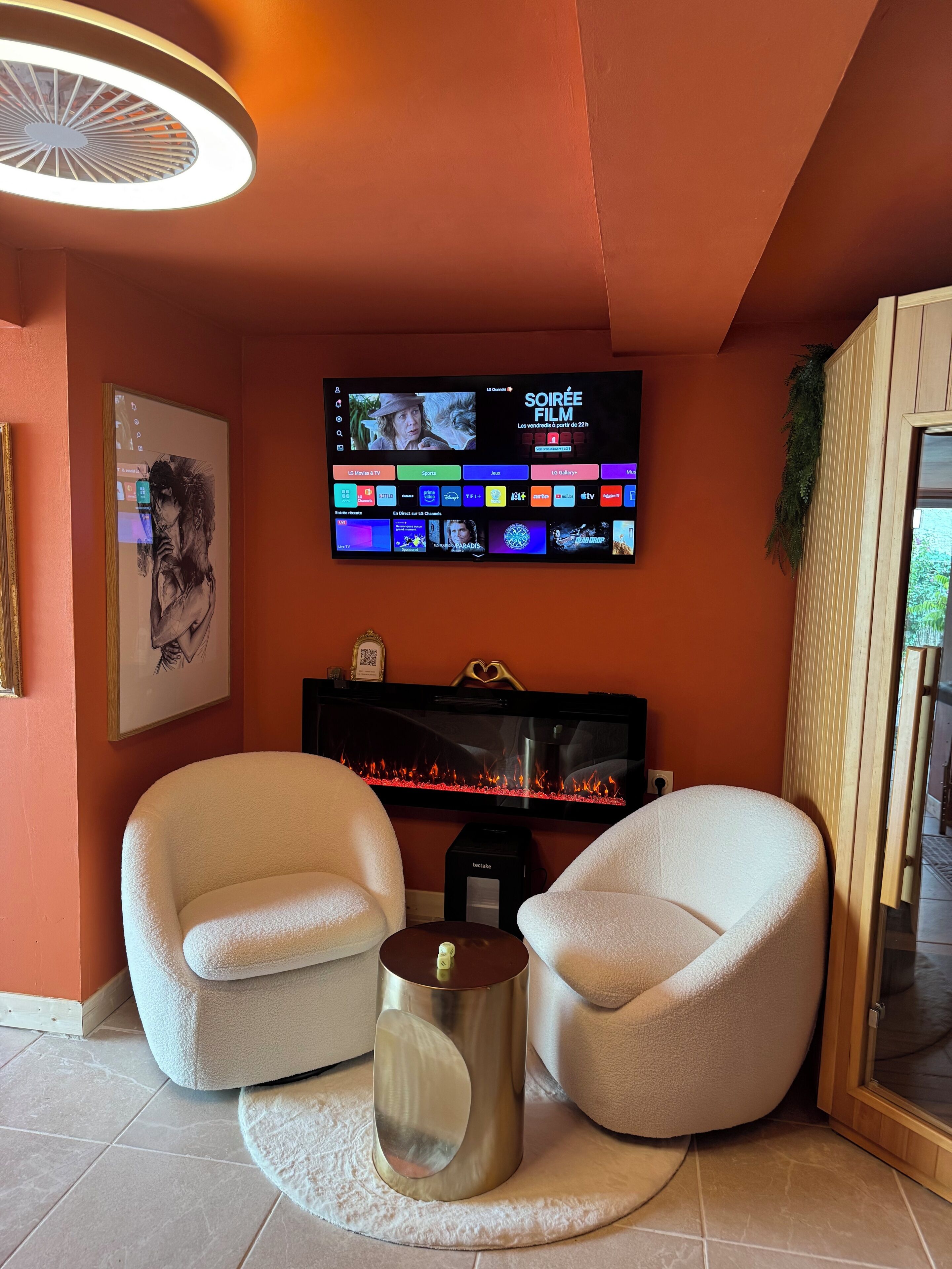 A living room with a flat-screen TV mounted on the wall, two white armchairs, a round coffee table, and a fireplace.