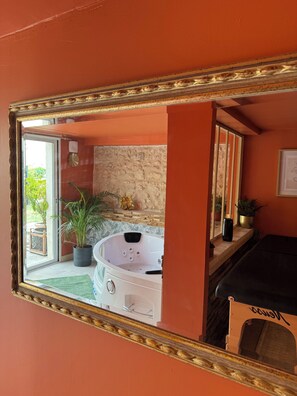A room with a large mirror reflecting a jacuzzi, a stone wall, and a window with a view of greenery.