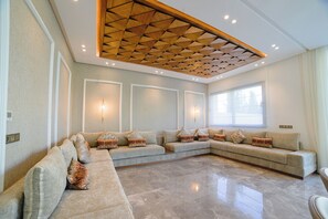 A spacious living room with a large L-shaped sectional sofa, a polished marble floor, and a geometric wooden ceiling design. - Villa Yasmine – Luxury villa with pools, hammam, and gym in Marrakech (Douar Moulay Abbas)